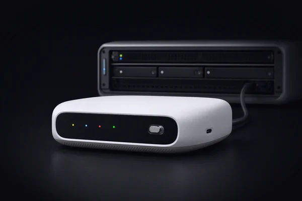A home compute node with four coloured lights and a sharing switch turned on, cabled to a blurred server rack behind it